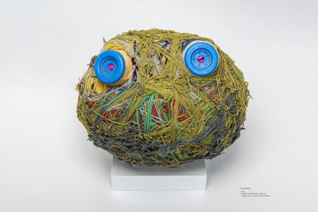 Fibers wound into an oval form, primarily in shades of olive green and gray, with layers of yellow, red, green, and gray beneath. Two bright blue, circular objects are embedded within the fibers, resembling wide, watchful eyes.