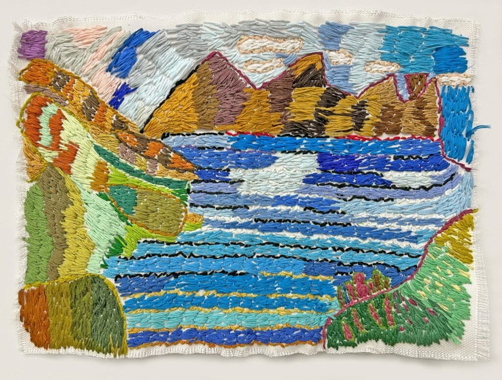 A multi-color thread based art piece showing the sea with a large mountain chain behind it. There are clouds in the sky above the mountains.